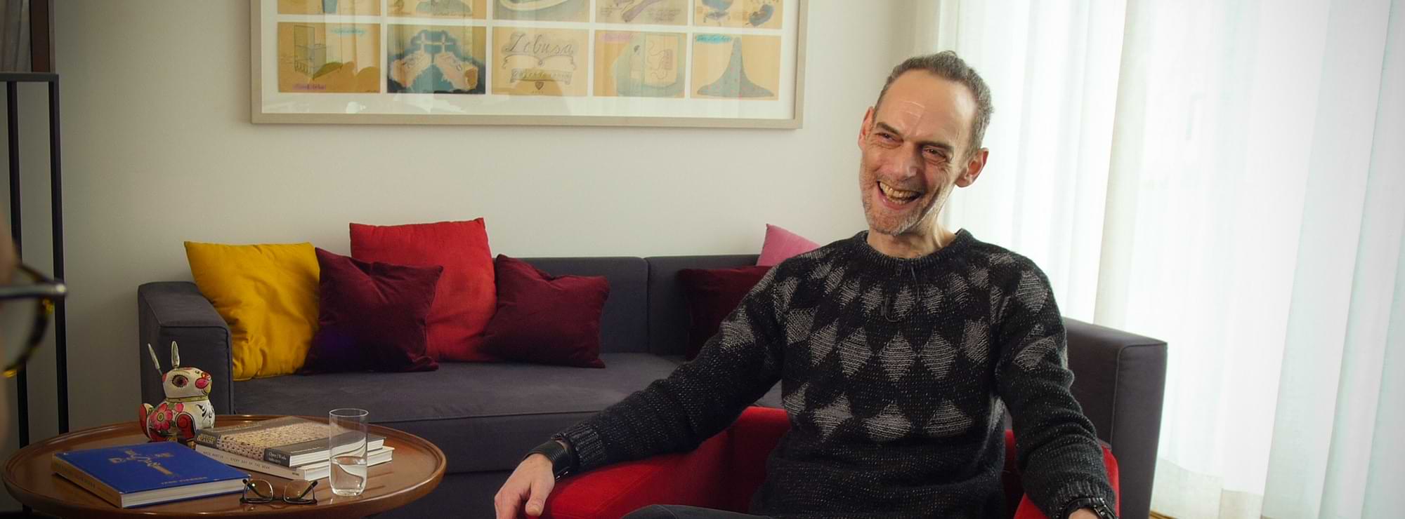 Arik Hayut in einem Wohnzimmer auf dem Roten Sessel, lachend