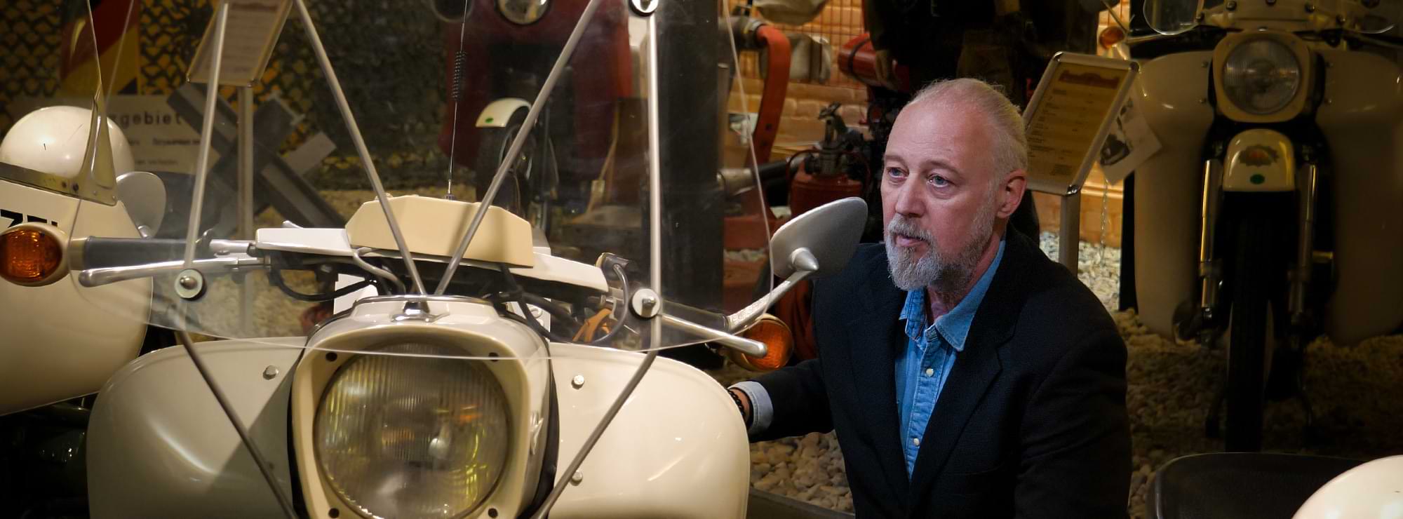 Thomas Müller in einem Motorradmuseum, knieend vor einer Maschine, und bewundert diese Thomas Müller in einem Motorradmuseum, knieend vor einer Maschine, und bewundert diese