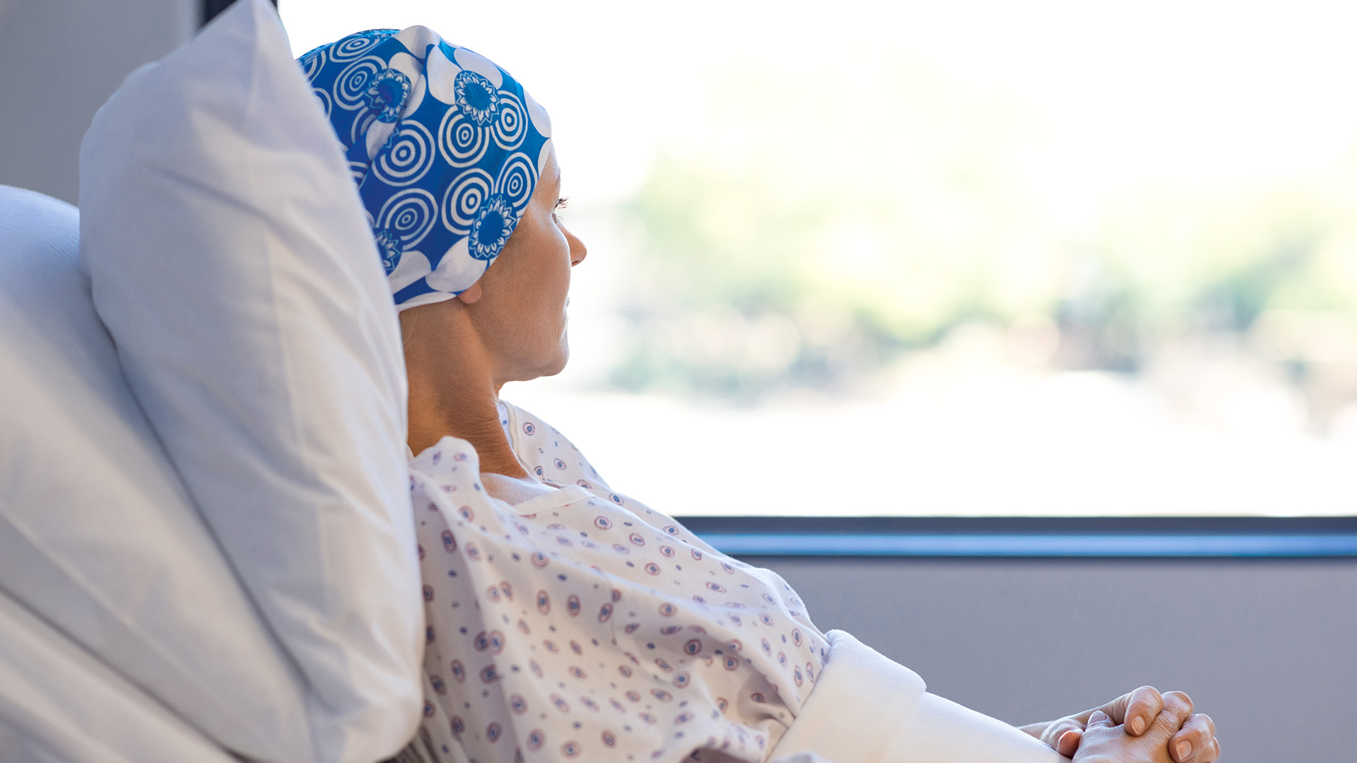 Frau mit Kopftuch nach Chemo schaut vom Krankenhausbett aus dem Fenster