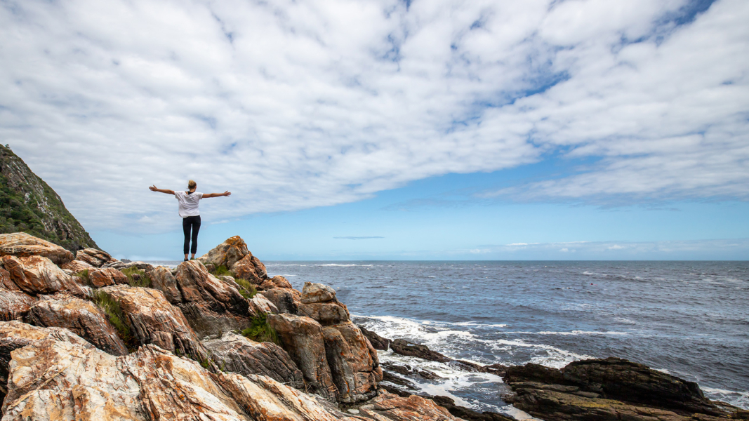 Person mit ausgestreckten Armen steht auf Felsen vor dem Meer