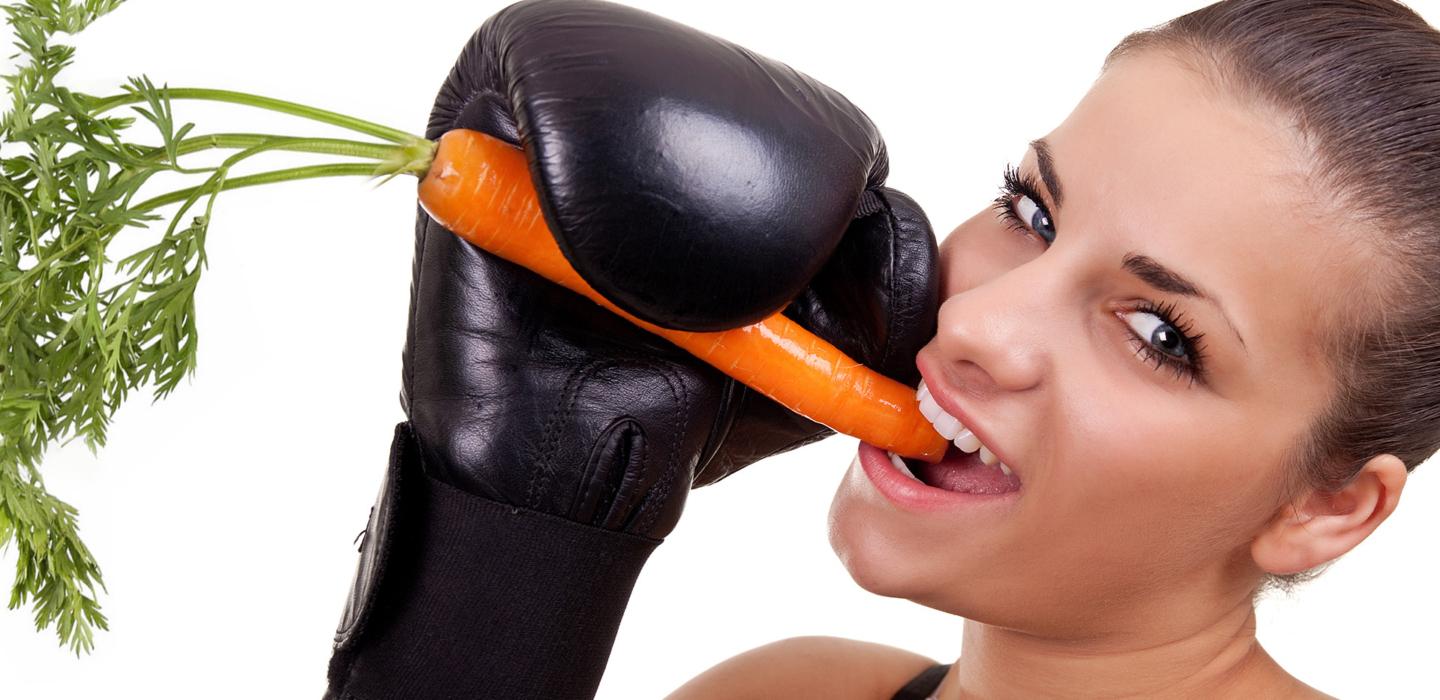 junge Frau mit Boxhandschuhen hat eine Möhre in der Hand und beißt ab, mit frechem Blick