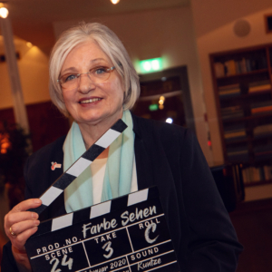 Anita Waldman mit einer Filmklappe in der Hand bei der Premierenveranstaltung "Farbe Sehen"