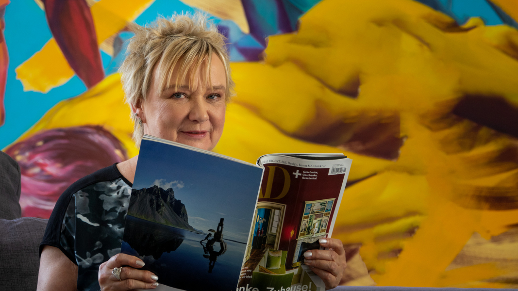 Sabine Schneider sitzt auf der Couch in einem Loft, hält ein Magazin in der Hand und schaut in die Kamera